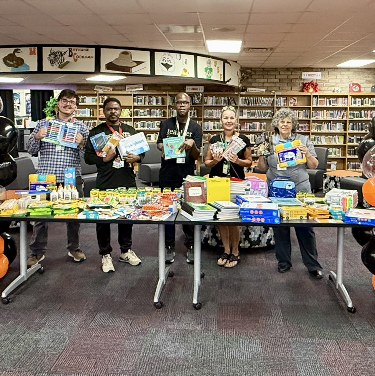 Picture of various teachers standing in front of a table, full of supplies with orange and black balloon display on either side