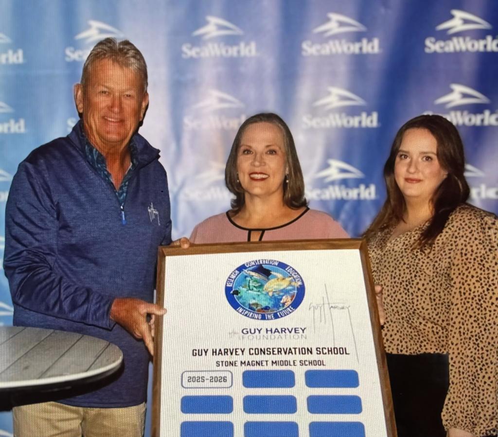 Dr. Alexander, Ms. Knowles, and Guy Harvey holding picture fram in front of seaworld banner