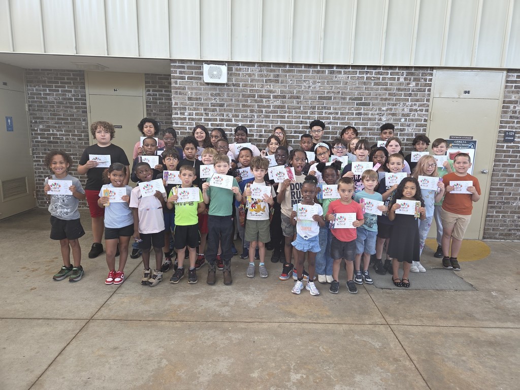 A large group of Columbia Elementary students standing together outside on the patio, proudly holding up their summer birthday certificates and small gift bag