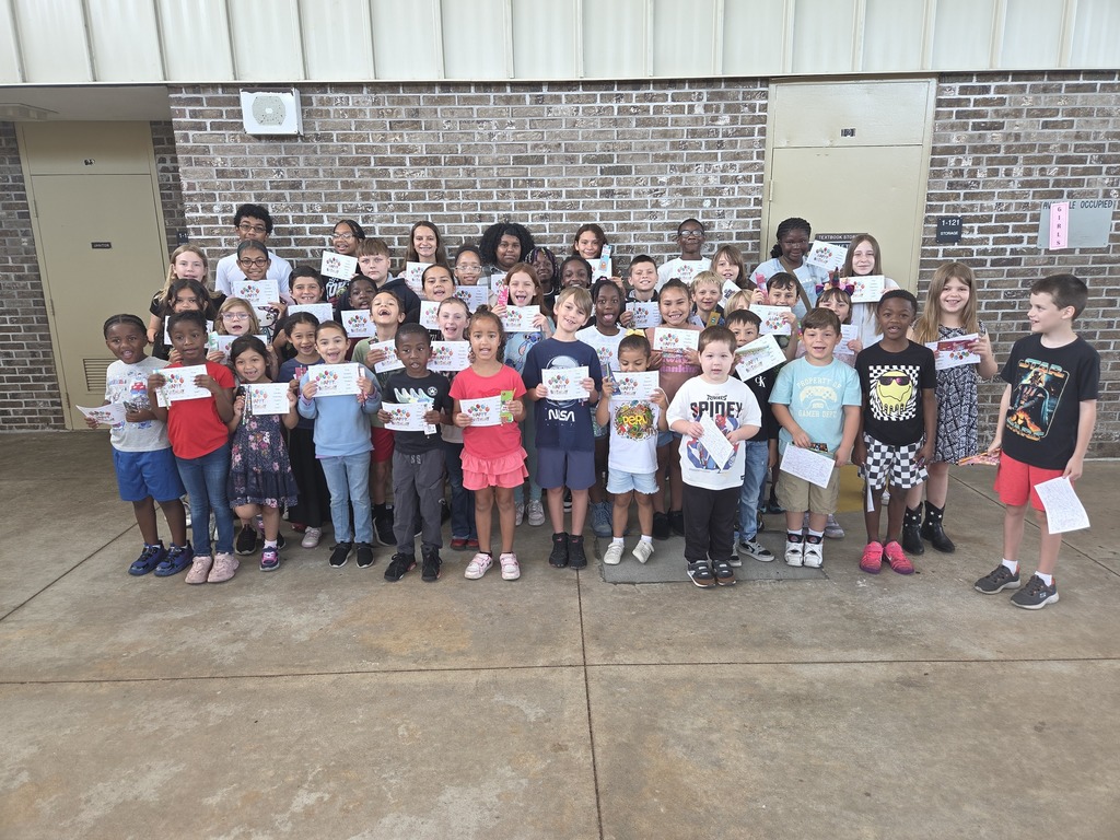 A large group of Columbia Elementary students standing together outside on the patio, proudly holding up their summer birthday certificates and small gift bag
