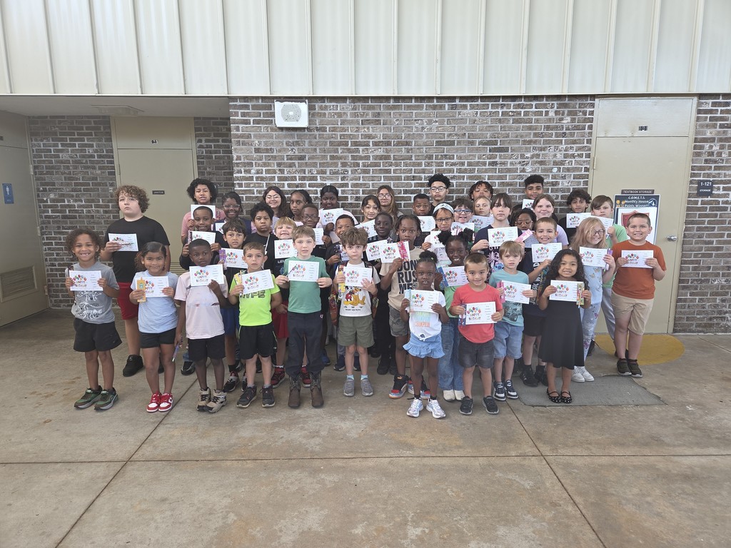 A large group of Columbia Elementary students standing together outside on the patio, proudly holding up their summer birthday certificates and small gift bag