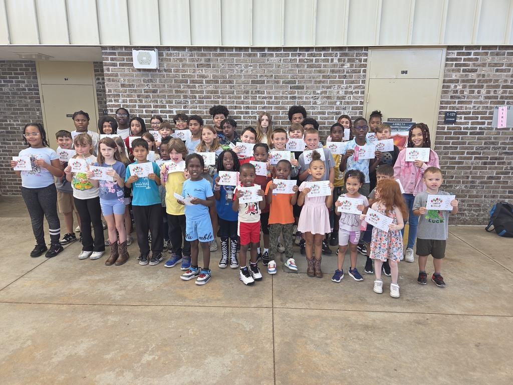 A large group of Columbia Elementary students standing together outside on the patio, proudly holding up their summer birthday certificates and small gift bag