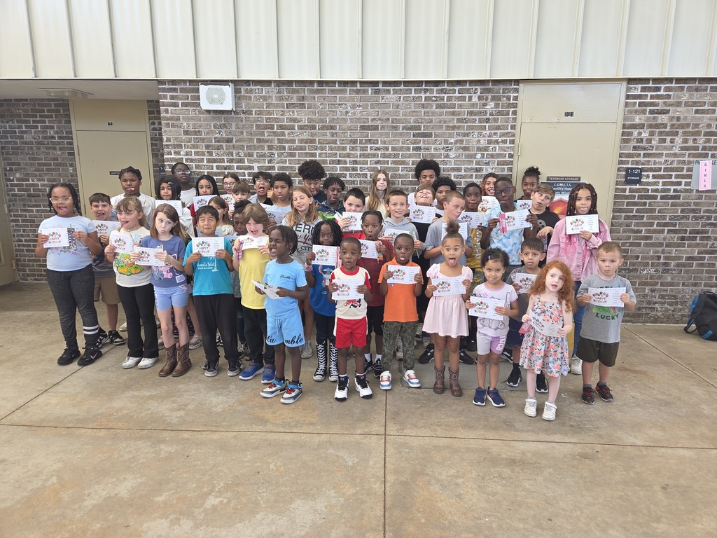 A large group of Columbia Elementary students standing together outside on the patio, proudly holding up their summer birthday certificates and small gift bag