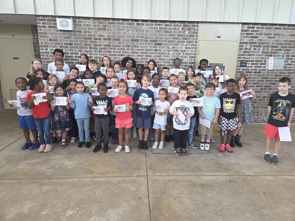 A large group of Columbia Elementary students standing together outside on the patio, proudly holding up their summer birthday certificates and small gift bag