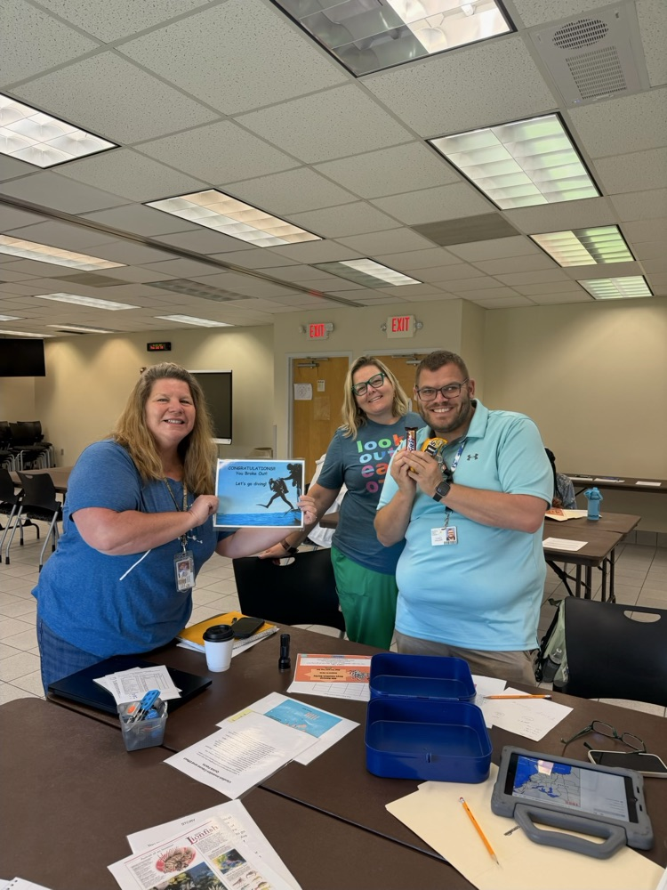 Three educators stand together in a traing room, smiling as they celebrate a completed activity. One participant holds a certificate that reads, Congratulations! You Broke Out! Let's go diving! While another holds candy prizes. Table with activity materials and classroom supplies are visible in the foreground. !