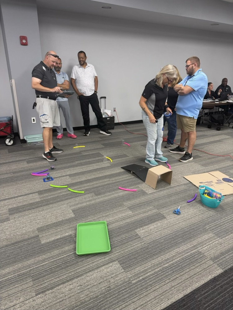Educators are participating in a Hands-on STEM challenge in a professional learning activity. Participants are standing around a course made with colorful plastic tubes, a binder as a ramp, and other materials.while collaborating , observing, and discussing the activity. Additional educators watch and work in the background.