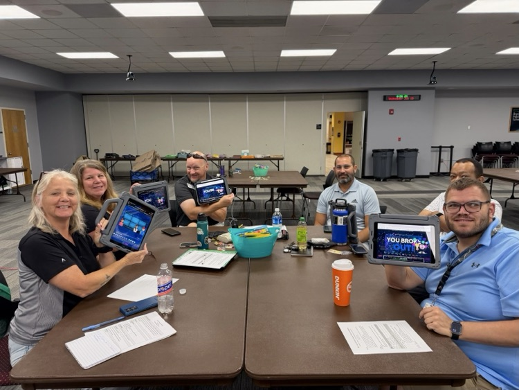 a group of educators are sitting around a table during a professional learning session, smiling, and holding up iPads displayingc ompleted digital escape room. Notebooks, drinks , and Workshop materials are spread across the table.