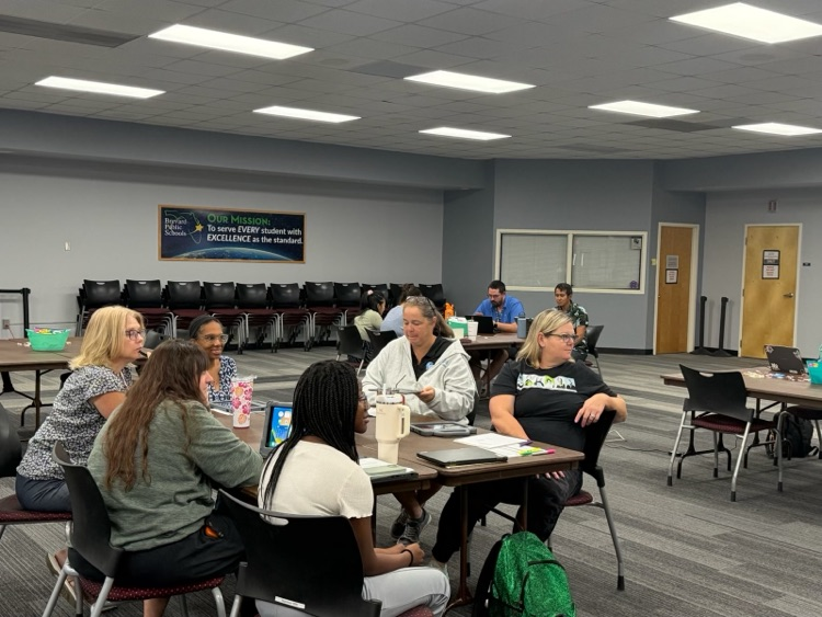 Educators sit around a table in a small group during a professional development session, listening to a presenter outside the frame. Laptops, ipads, notebooks, and other workshop materials.Additional participants work at a table in the the background  throughout the training room. A Brevard Public Schools mission statement is displayed on the wall in the background.Mission Statement 