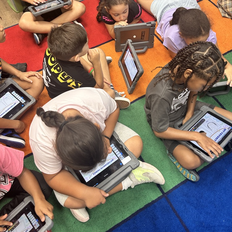 a group of students sitting and lying on a carpet with their iPads working on the “All About Me” lesson.