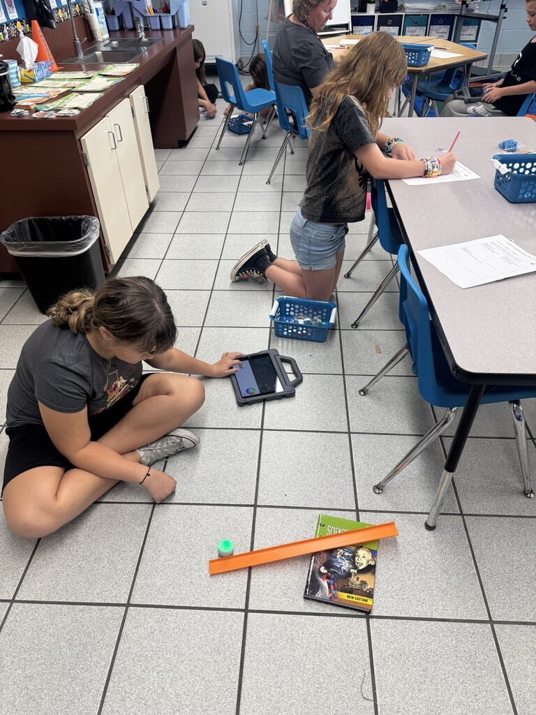 Two sixth-grade students completing a force and motion activity by building a ramp on the floor with a textbook, orange plastic track, and a Sphero Mini. One student is controlling the Sphero Mini on an iPad, the other student records observation on paper.