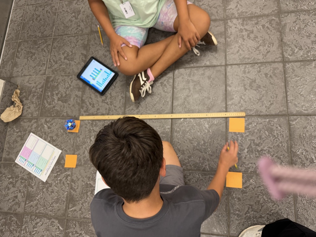 Two fifth- grade students using sticky notes, a yard stick, and a Sphero Bolt to calculate the perimeter and area of the rectangle.