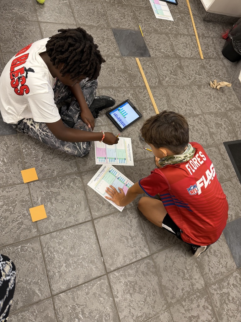 Two fifth- grade students sitting on the floor, recoding their data on paper after finding the perimeter and area of the rectangle.