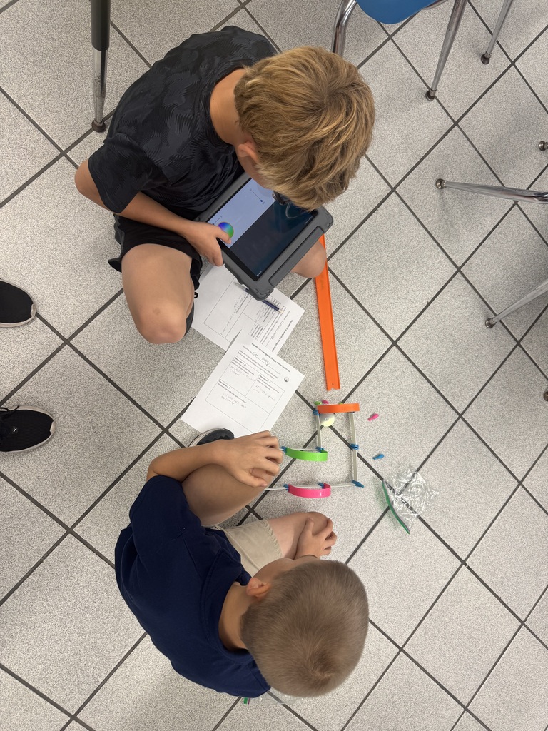 Two sixth- graders sitting on the floor with their ramp they built with an orange plastic track, paper arches, paper walls and tape. One student is controlling the Sphero Mini with an iPad. The other student is observing.