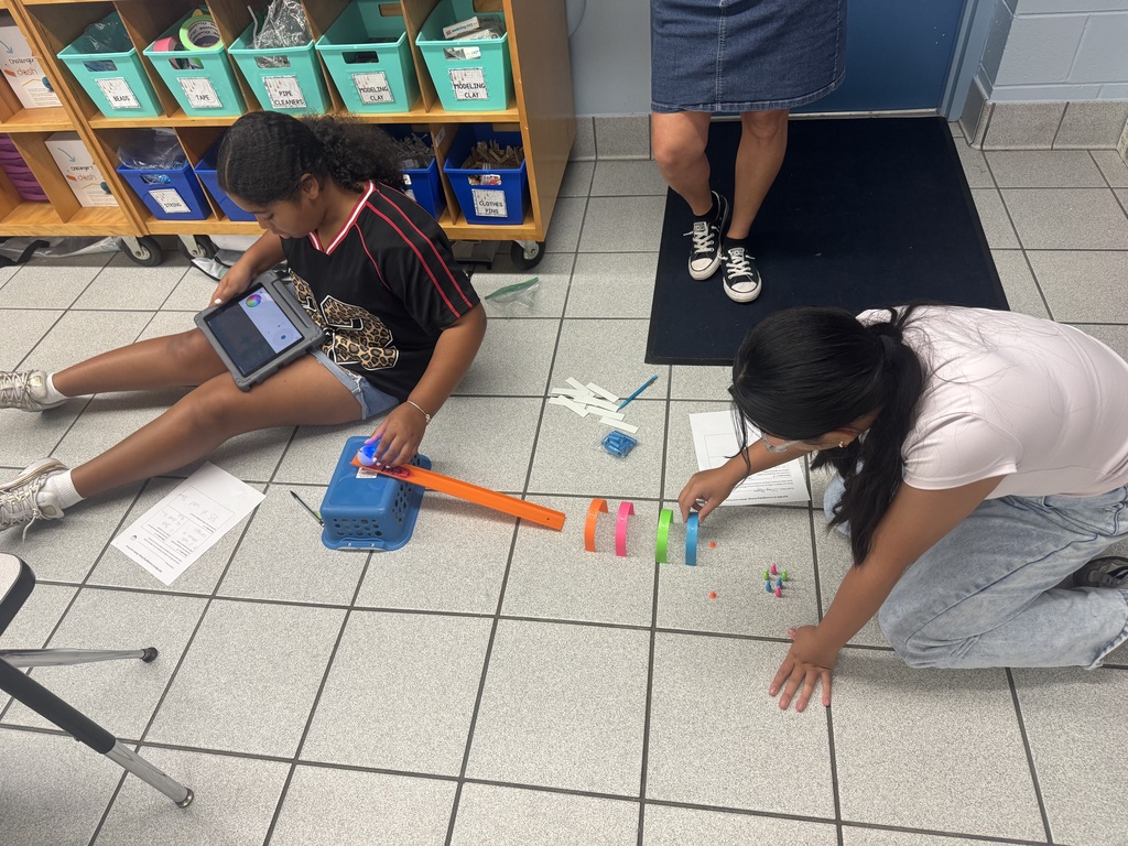 Two sixth- graders sitting on the floor building a ramp and tunnel using a small blue basket, orange plastic track and paper arches. One student is placing the Sphero mini at the top of the track while holding an iPad. The other student is placing the paper arches.