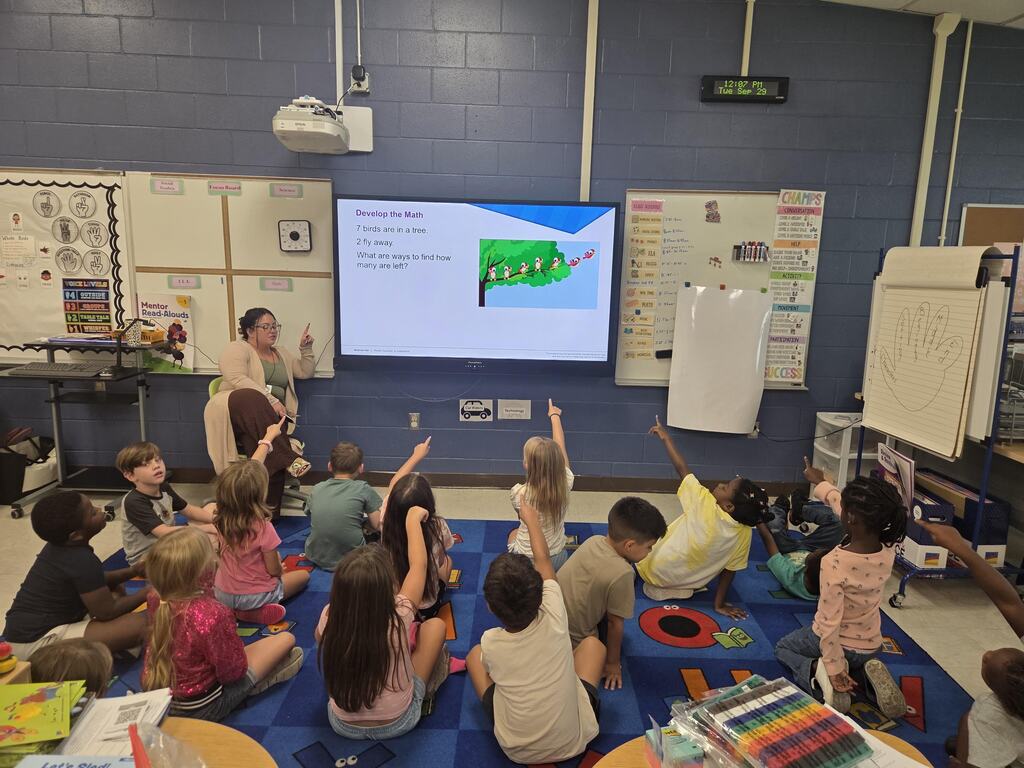 A teacher sitting next to a digital display showing a math word problem, while a group of elementary students sitting on a colorful carpet raise their hands to answer.