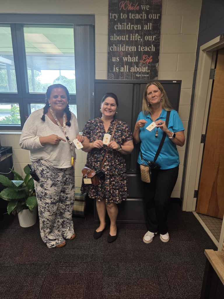 Three women smiling and holding up small white emergency panic buttons in an office setting, with an inspirational quote on the wall behind them