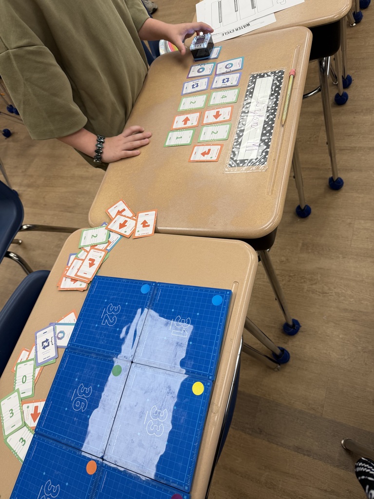 Students collaborate at grouped desks, arranging coding cards and testing a KaiBot sequence to model the movement of a water droplet through the water cycle. A blue activity mat is set up nearby for robot navigation.