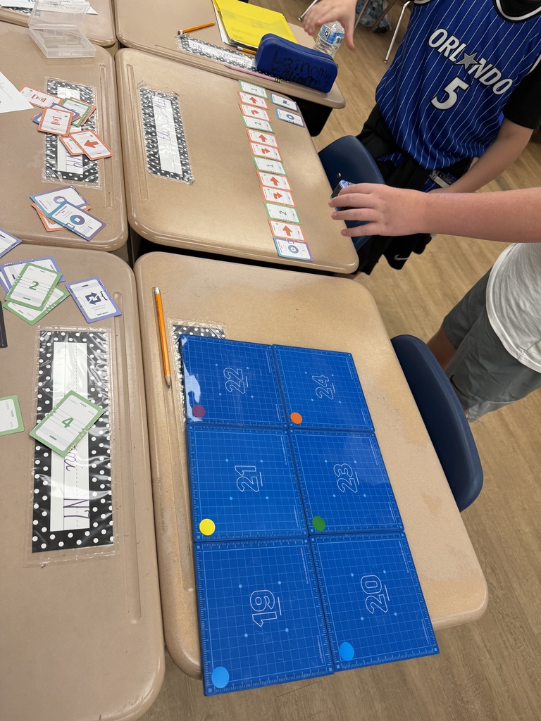 Fifth-grade student uses a KaiBot and coding cards to program a path representing stages of the water cycle. A blue grid mat and movement cards are arranged on classroom desks as part of a hands-on STEM activity.