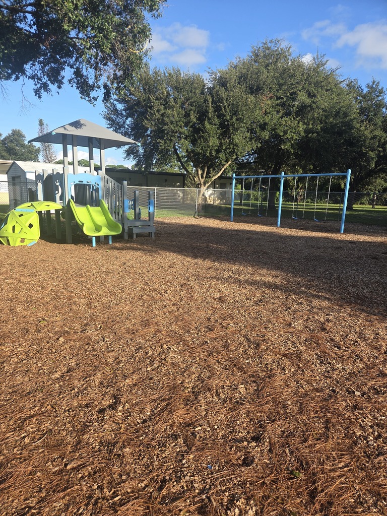 A newly installed outdoor playground at Columbia Elementary School featuring bright turquoise, lime green, and purple climbing structures, monkey bars, and balancing steps. The playground is enclosed in a black border filled with fresh wood chips, set against a large grassy field and a blue sky with scattered clouds.