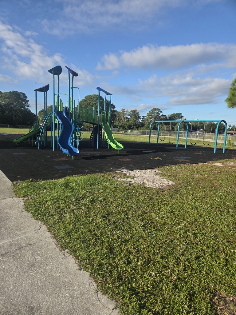 A playground at Columbia Elementary School with a small gray and blue climbing structure featuring double lime green slides and a green play dome, next to a blue swing set. The play area is covered in fresh wood chips and shaded by mature oak trees, with a school building and fence in the background under a blue sky.