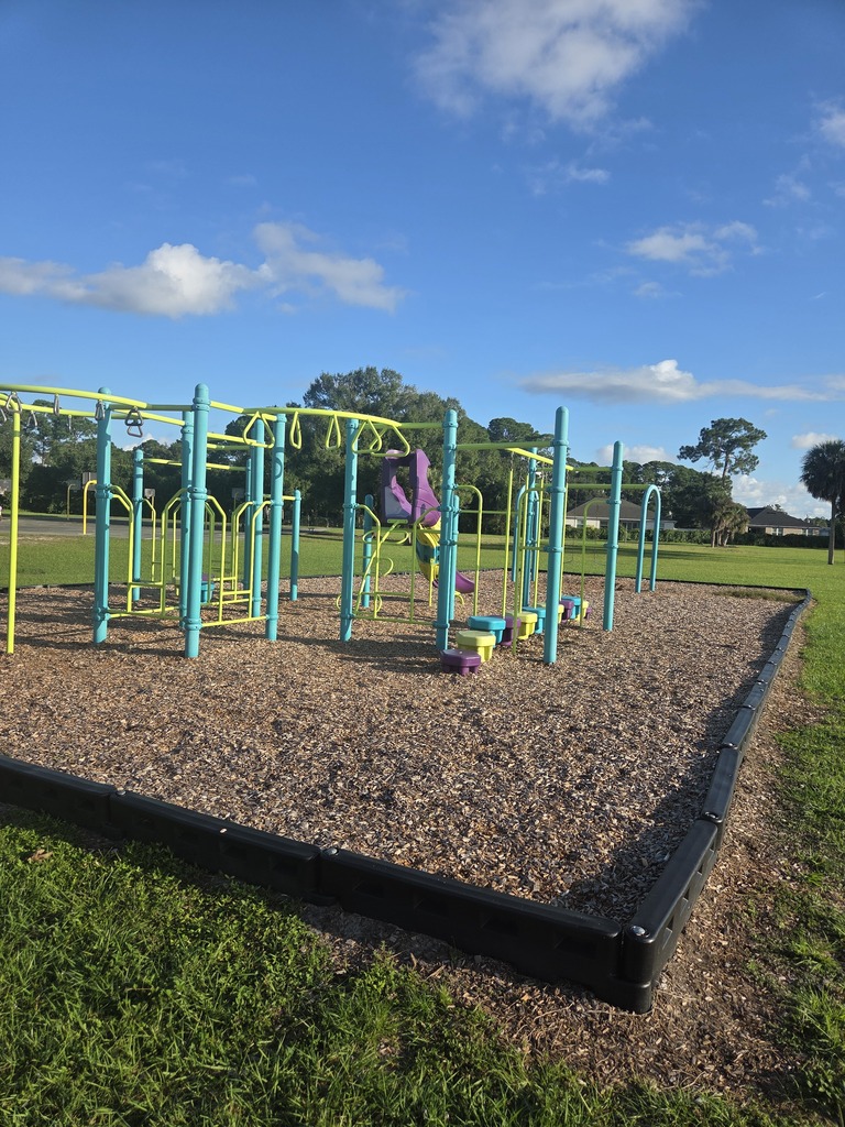 A colorful playground at Columbia Elementary School featuring a large blue, green, and purple play structure with multiple slides and climbing features, alongside a blue swing set. The equipment sits on a black protective safety matting next to an open grassy field and a concrete walkway under a partly cloudy sky.