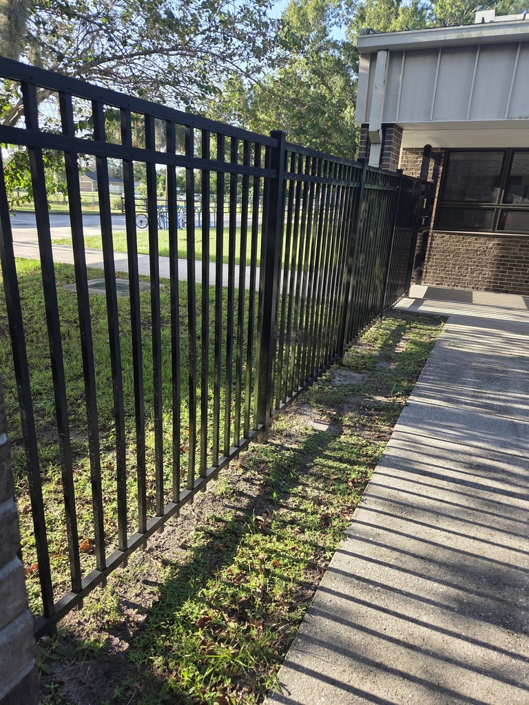 A tall, black metal security fence installed alongside a concrete walkway on a school campus, highlighting recent facility upgrades.