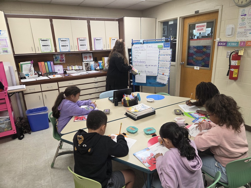Mrs. Sutfin, a 2nd grade reading interventionist, stands at a whiteboard teaching a lesson to a small group of students working at a table in a classroom.