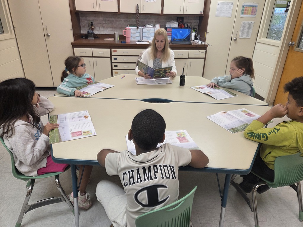 Mrs. Good, a 3rd grade reading interventionist, reads a book aloud to a small group of students seated around a horseshoe table in a classroom.