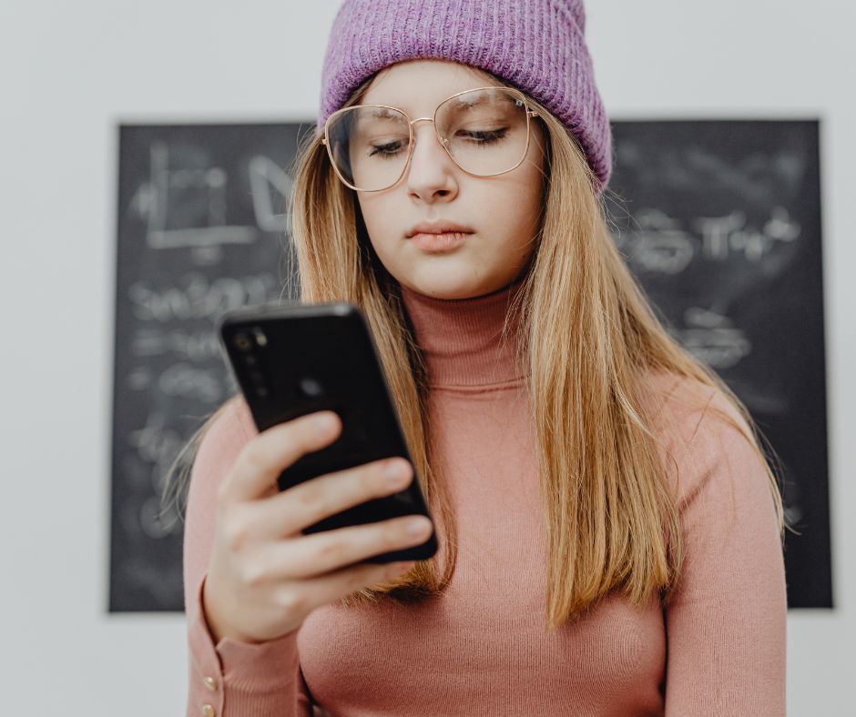 The image is of a middle school aged female student wearing glasses and a knit beanie hat. The student is looking at a black cell phone in her hand.