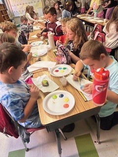 Elementary students sit at desks painting rocks. Each student has a paper plate with colorful paint and brushes.