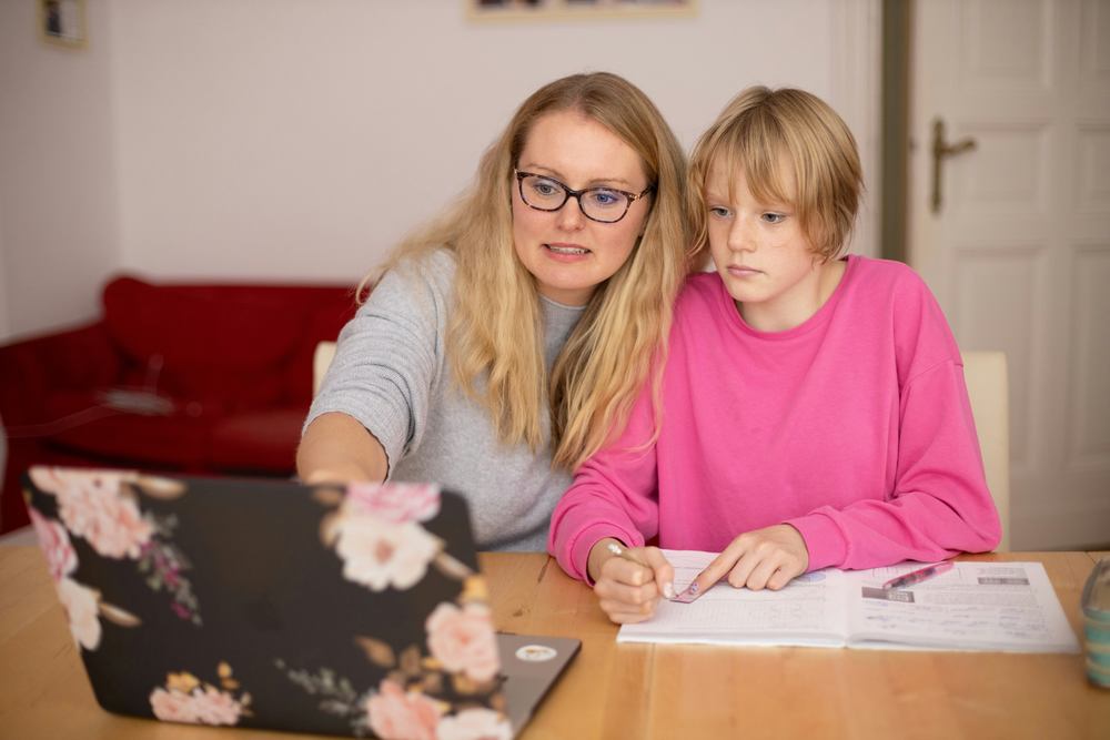 This image is of an adult with long blonde hair and plastic frame glasses helping a middle school boy with blonde hair. They appear to be working on school work. The woman has a laptop open on the table, and the middle school boy has a notebook open to write and is looking at the laptop.