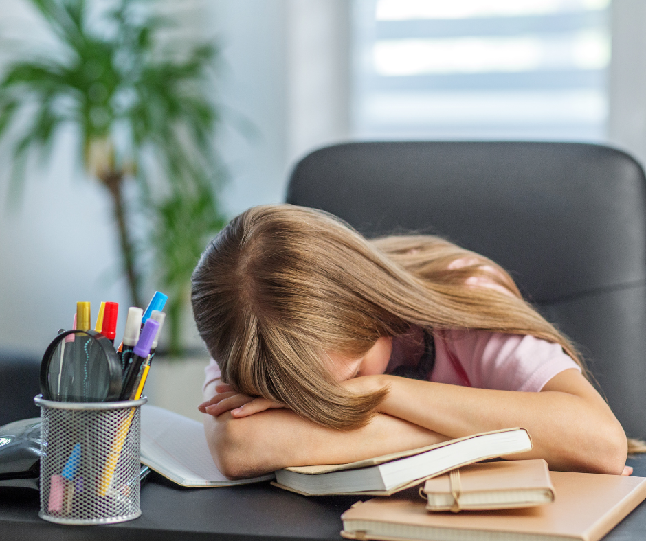 The image is a middle school age female student sitting in a black desk chair. She has her head down on top of her arms and her arms crossed on top of the desk. There are books, notebooks, and a pencil cup with pens and pencils. The student appears frustrated.