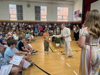 A student in a tan outfit stands in the center of a school gymnasium floor receiving an award from a man in a beige suit, while a girl in a striped dress stands nearby. A large audience of students and families fills the bleachers and chairs in the background.