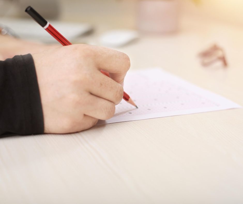 The image is an up-close look of a student hand holding a pencil and appearing to bubble in an answer sheet for a test.