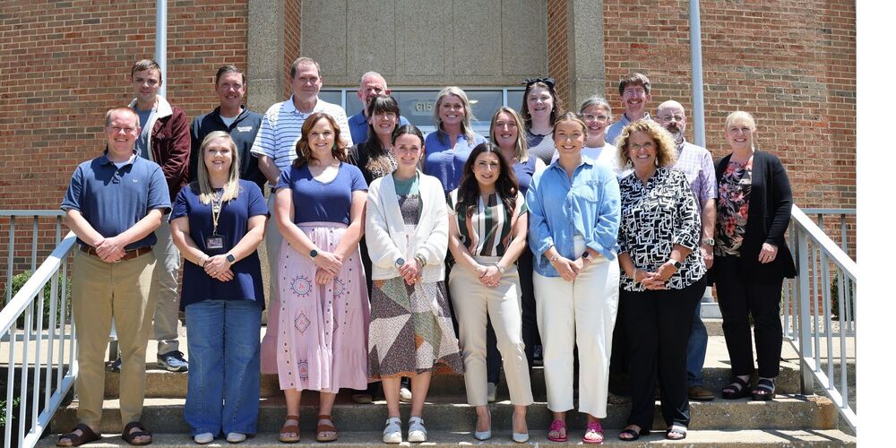 A group of approximately 17 participants in the Bristol Tennessee City Schools Aspiring Leaders Academy stands on the front steps of a brick school building on a sunny day. The group is arranged in three rows and smiles toward the camera. Participants wear a mix of professional and business-casual attire, including dresses, cardigans, polos, and button-down shirts. The photo was taken outside the building entrance, with brick walls, handrails, and the school's doorway visible in the background.