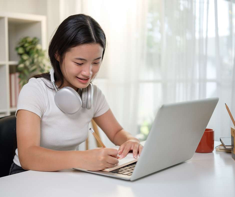Image is a middle school aged female student with dark hair looking down at a laptop computer. The student appears to be writing with a pencil and working on the laptop and is wearing headphones around her neck.