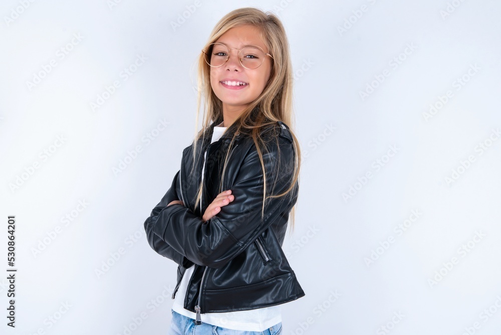 This photo is of a middle school age girl wearing biker jacket and glasses over white background standing with folded arms and smiling