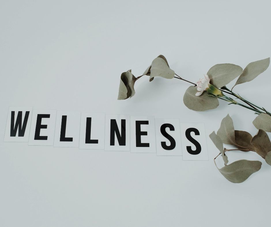 The image has individual black capital letters spelling out the word WELLNESS. There is a small bunch of leaves and a flower to the right and above the word.
