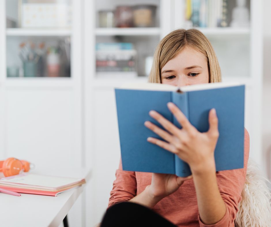 The image is of a middle school aged girl with dark blonde hair reading a novel seemingly at home. The book is open in front of the student covering part of her face.