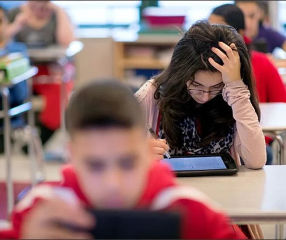 The image features a middle school aged female student with her elbow resting on a student desk and her head in her hand. She appears to be working on a tablet. Other students are blurred in the background around her working on an individual device as well. 