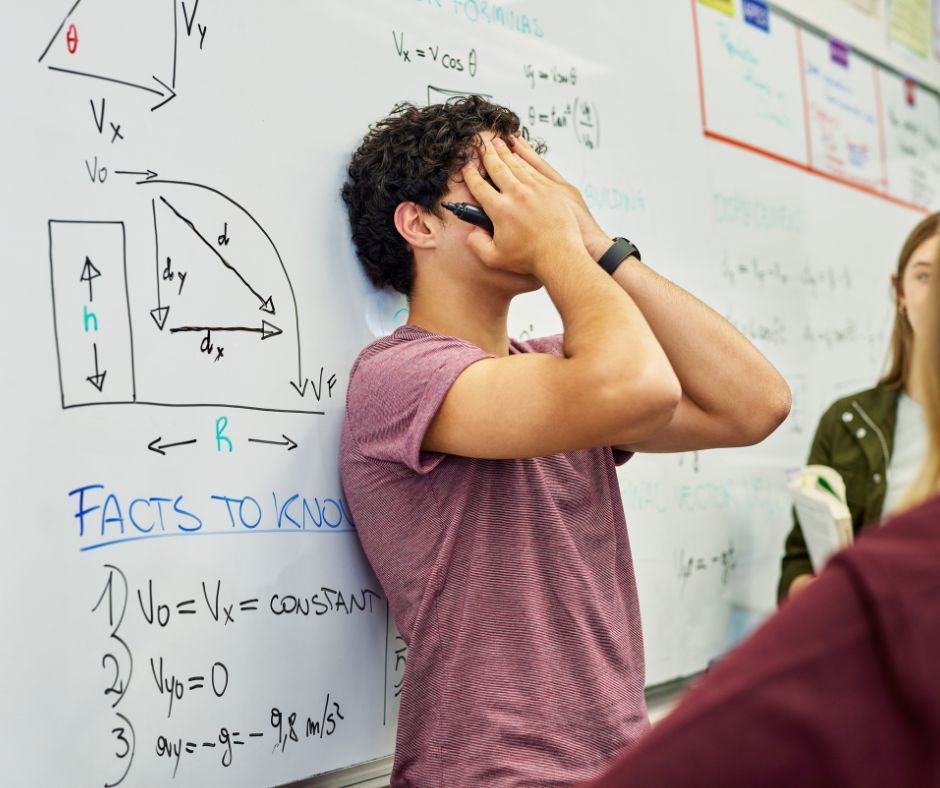 The image is of a male student leaning his back against a dry erase board in a classroom. He is holding a dry erase marker with his head in his hands appearing frustrated or confused as if he is working a problem on the board in front of the class.