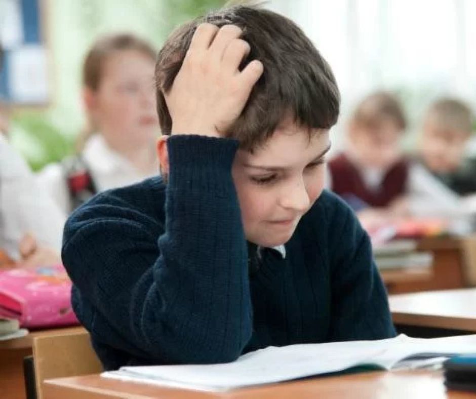 The image is of a middle school aged boy sitting at a test. He has his elbow on the desk and his hand on top of his head as if he is concentrating. He has papers spread out on the desk in front of him.