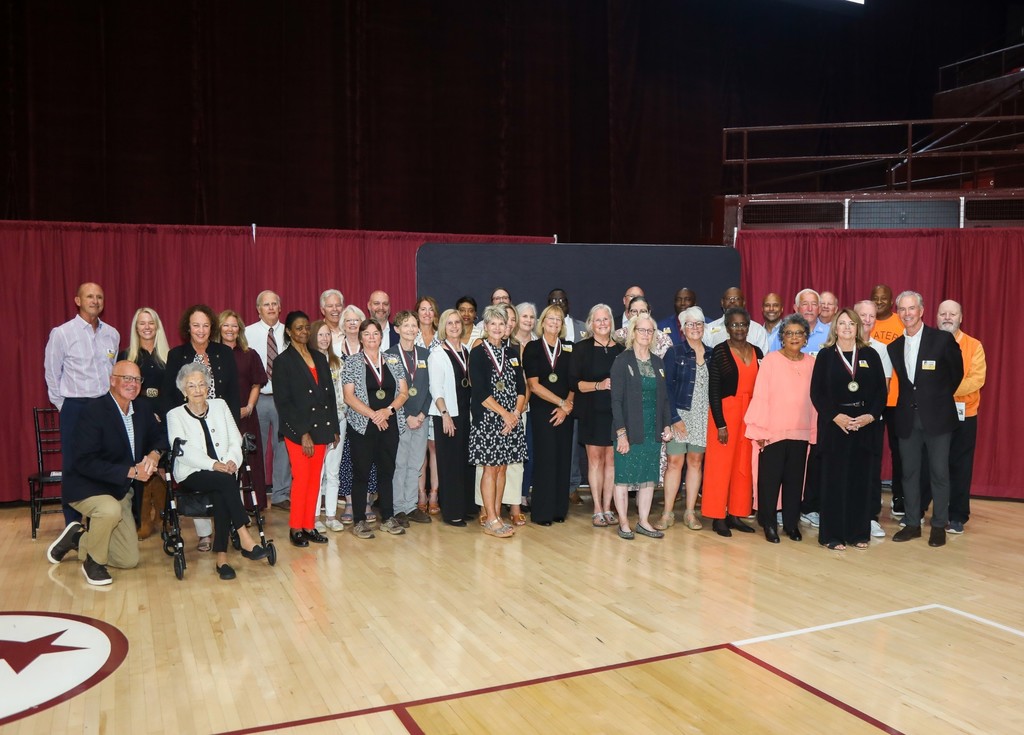 A large group of Bristol Tennessee City Schools Athletic Hall of Fame inductees pose together in a gymnasium. The group includes men and women of various ages, some wearing medals around their necks, with red curtains and a maroon-and-white floor visible in the background.