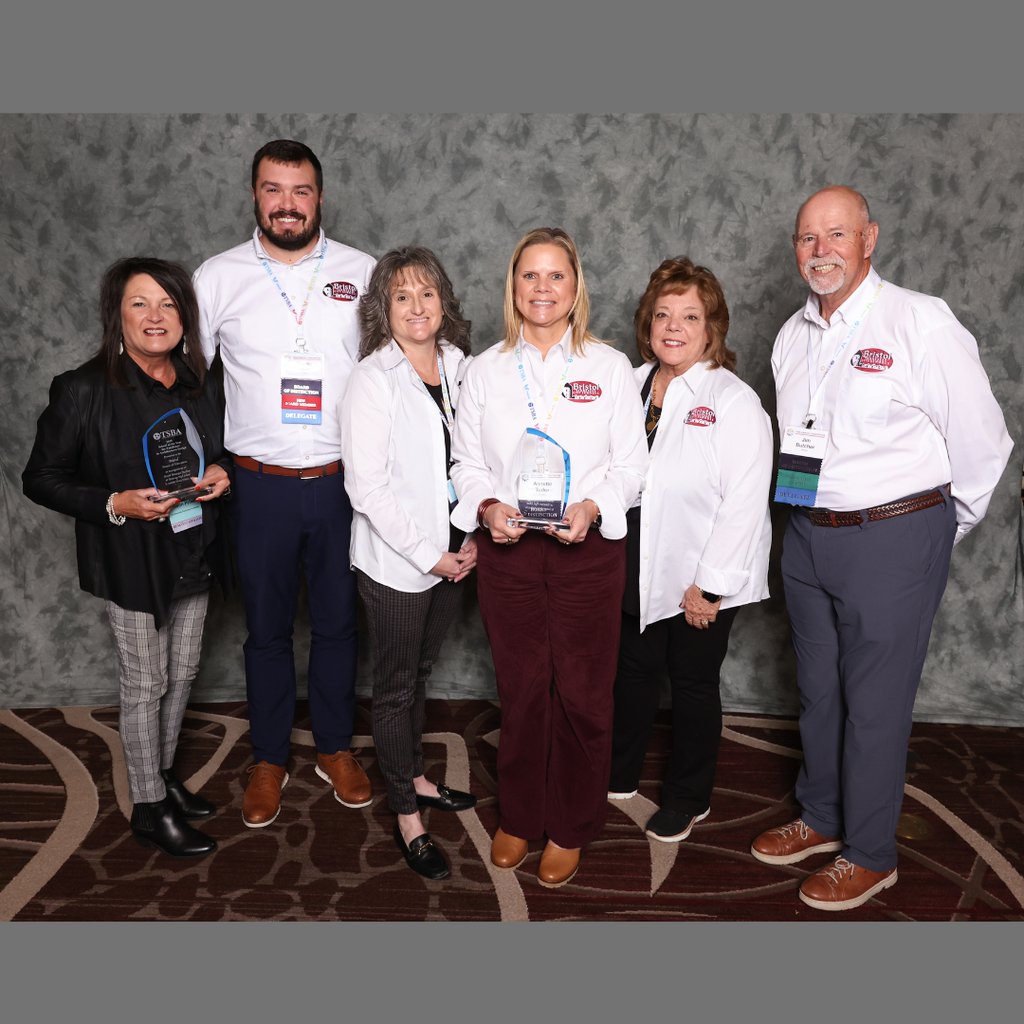 A group of six adults stands together indoors in front of a gray backdrop, smiling for a photo. Two people hold glass TSBA awards. Several individuals wear white shirts with the Bristol Tennessee City Schools logo, while others are dressed in professional attire. They appear to be attending a conference or awards event, as shown by their name badges and lanyards.