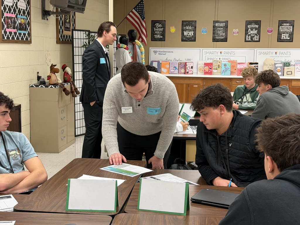 A Regions Bank volunteer leans over a student’s desk, helping him work through his budgeting worksheet during the Reality Check simulation.