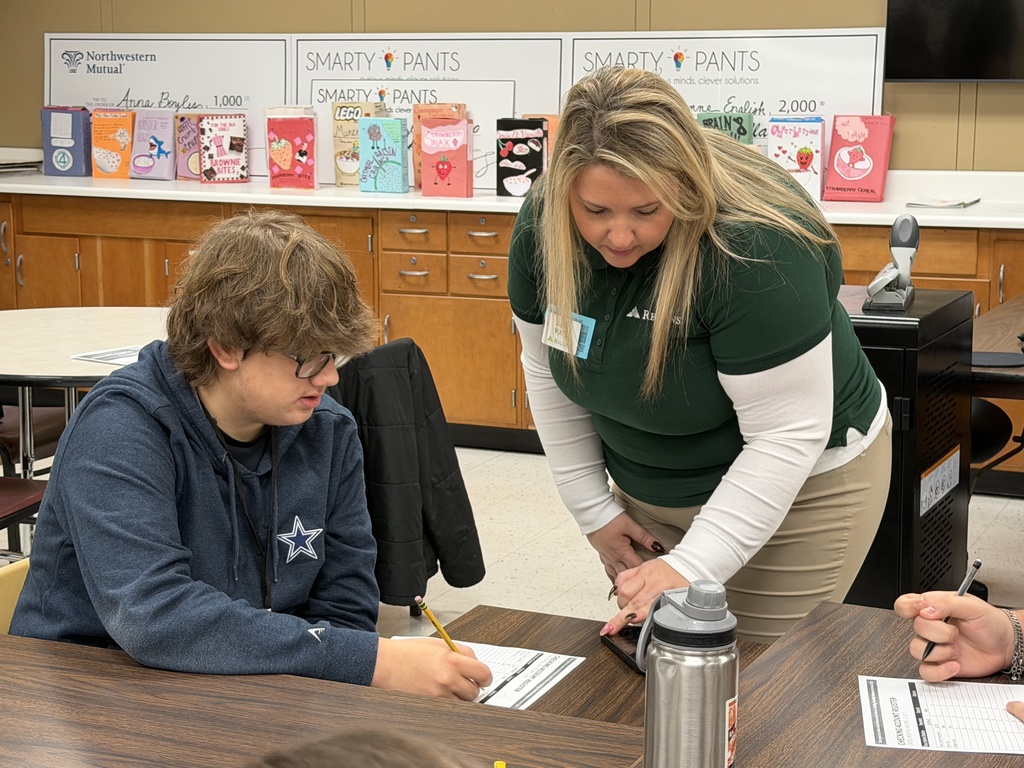Two Regions Bank volunteers assist students at separate tables as they calculate expenses; students are completing worksheets for the Reality Check budgeting lesson.