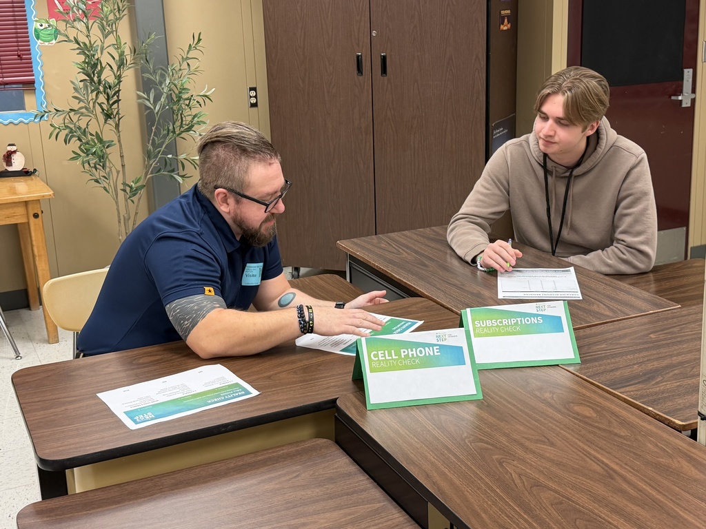 A male Regions Bank volunteer in a suit speaks with a group of students as they complete their transportation and food budgeting sheets.