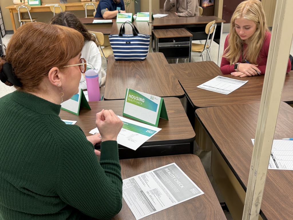 A Regions Bank volunteer in a green sweater discusses housing budget options with a student who is reviewing the Reality Check worksheet.
