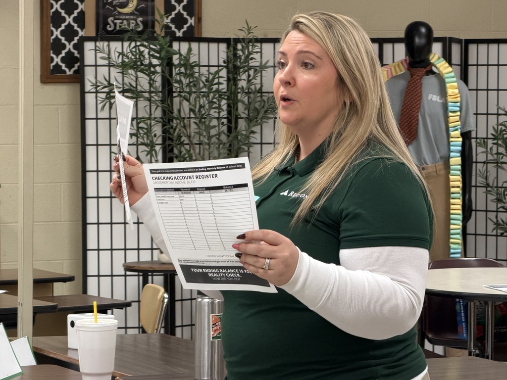 A set of table tents labeled “Utilities” and “Housing” sits on a classroom table alongside printed Reality Check worksheets used in the budgeting simulation.