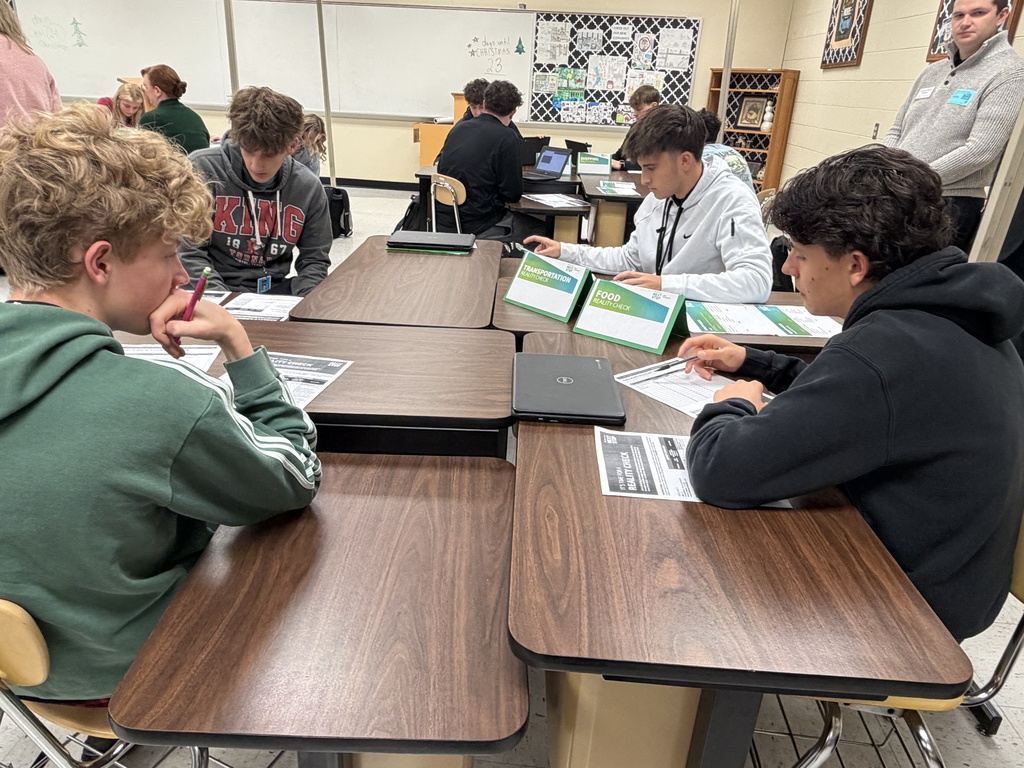 A classroom of high school students sits at grouped tables while five Regions Bank volunteers stand at the front of the room introducing the Reality Check budgeting activity.